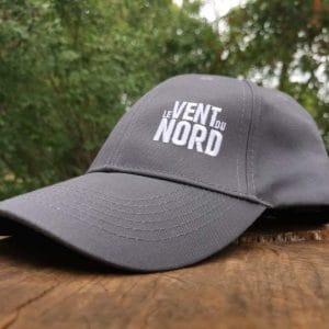 Une casquette de baseball grise avec le texte "le vent du nord" brodé dessus, posée sur une surface en bois avec de la verdure en fond.